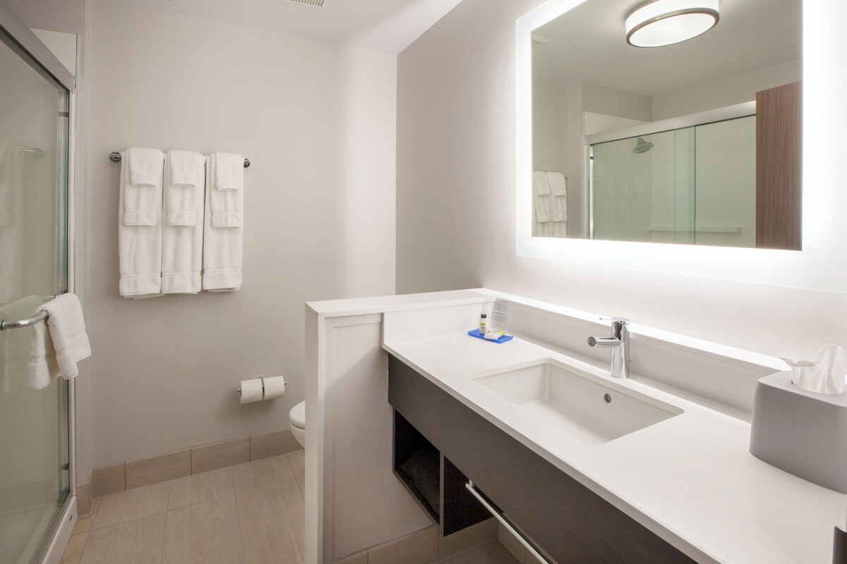 A modern bathroom with a glass shower enclosure, a white sink with a mirror above it, and three towels hanging on the wall.