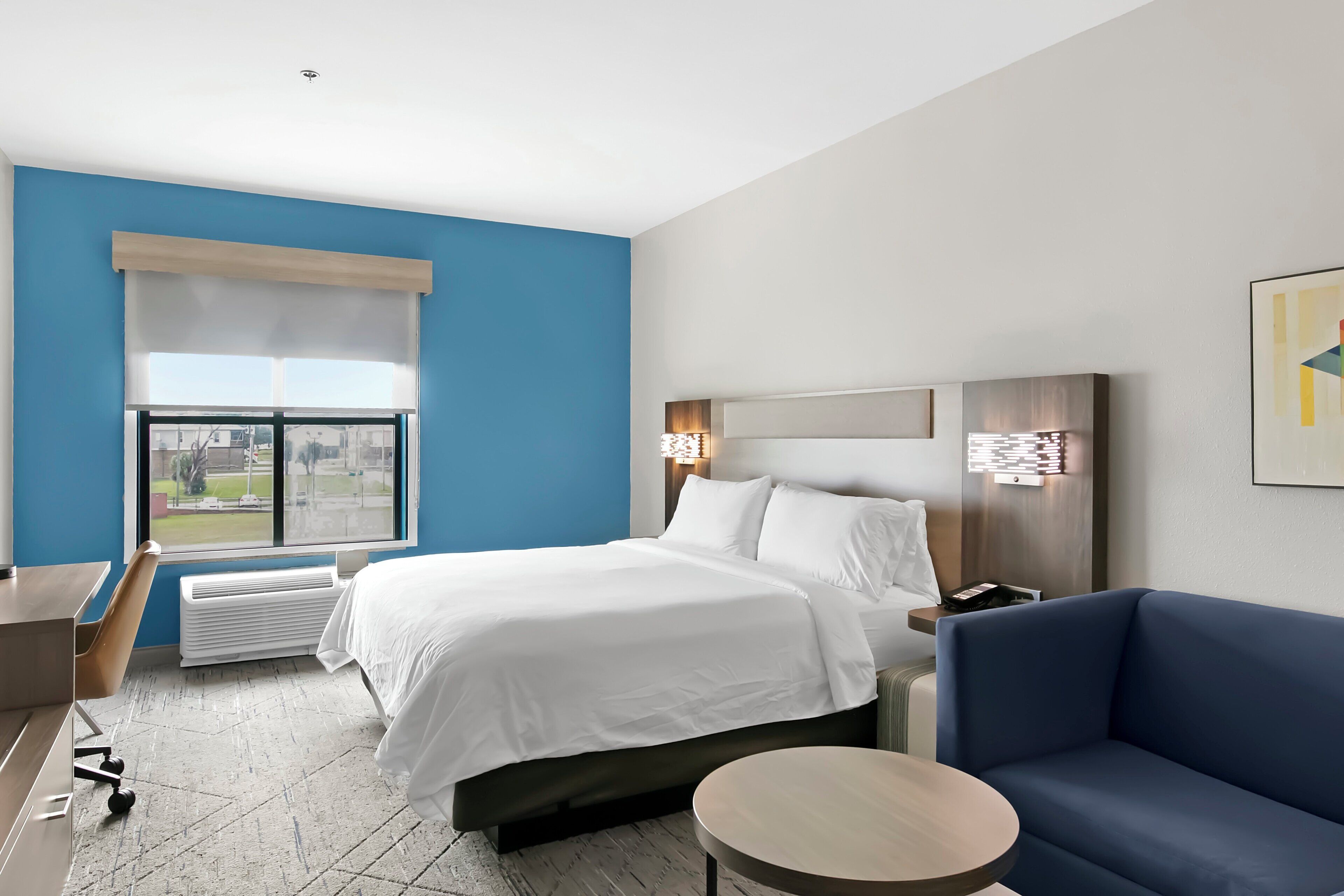 Ett hotellrum med en säng, ett skrivbord, en stol, en blå soffa, ett runt bord och ett fönster med utsikt över omgivningarna.