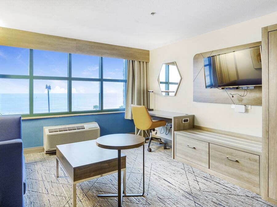 A hotel room with a large window, a desk, a chair, a TV, and a bed.