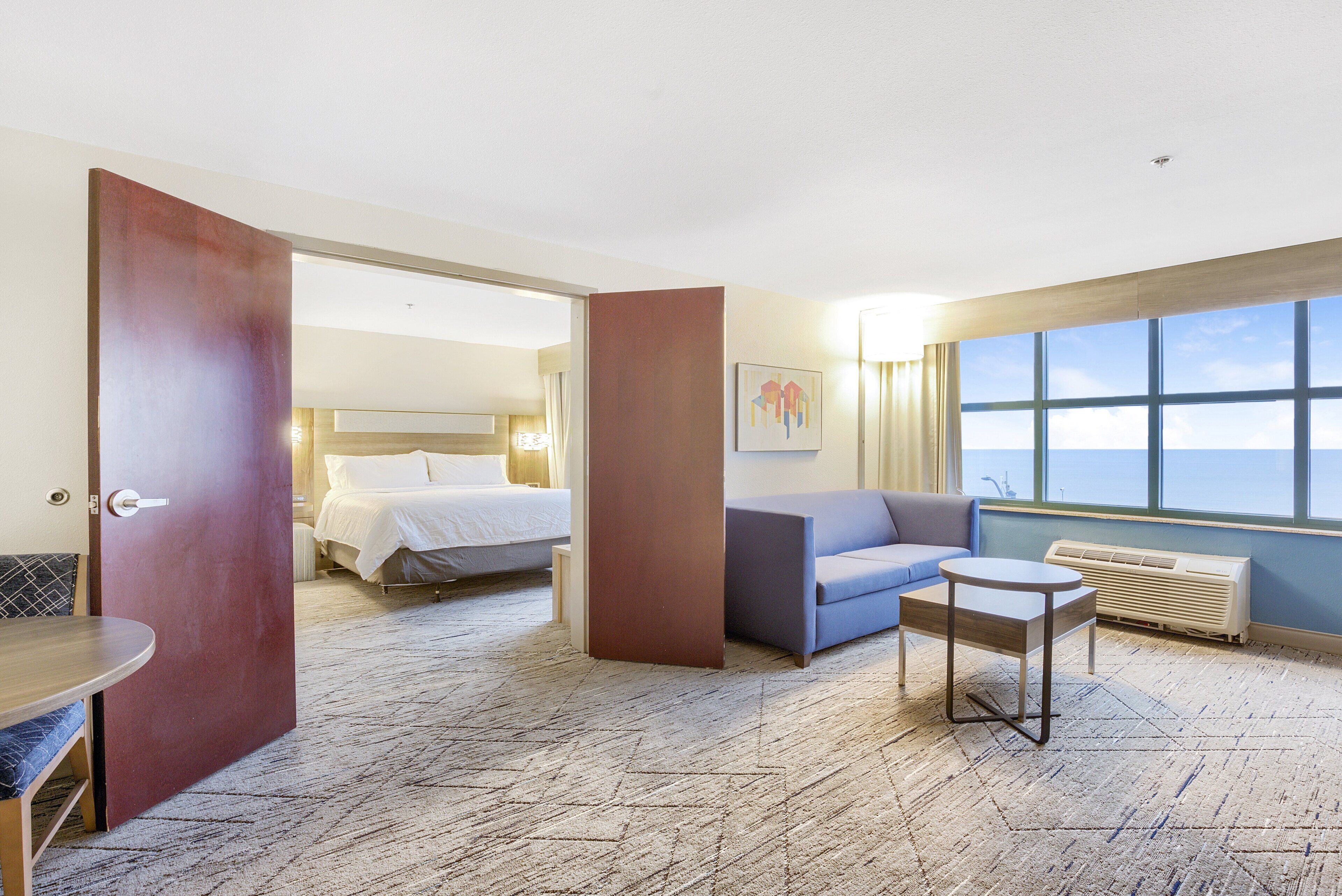 A hotel room with a bed, a sofa, a small table, and a view of the sky through large windows.