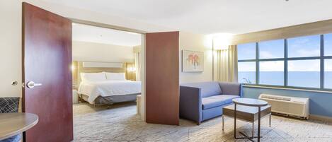 A hotel room with a bed, a sofa, a small table, and a view of the sky through large windows.