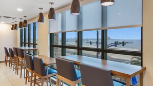 A dining area with tables and chairs, large windows with a view of the beach and traffic lights, and pendant lights.