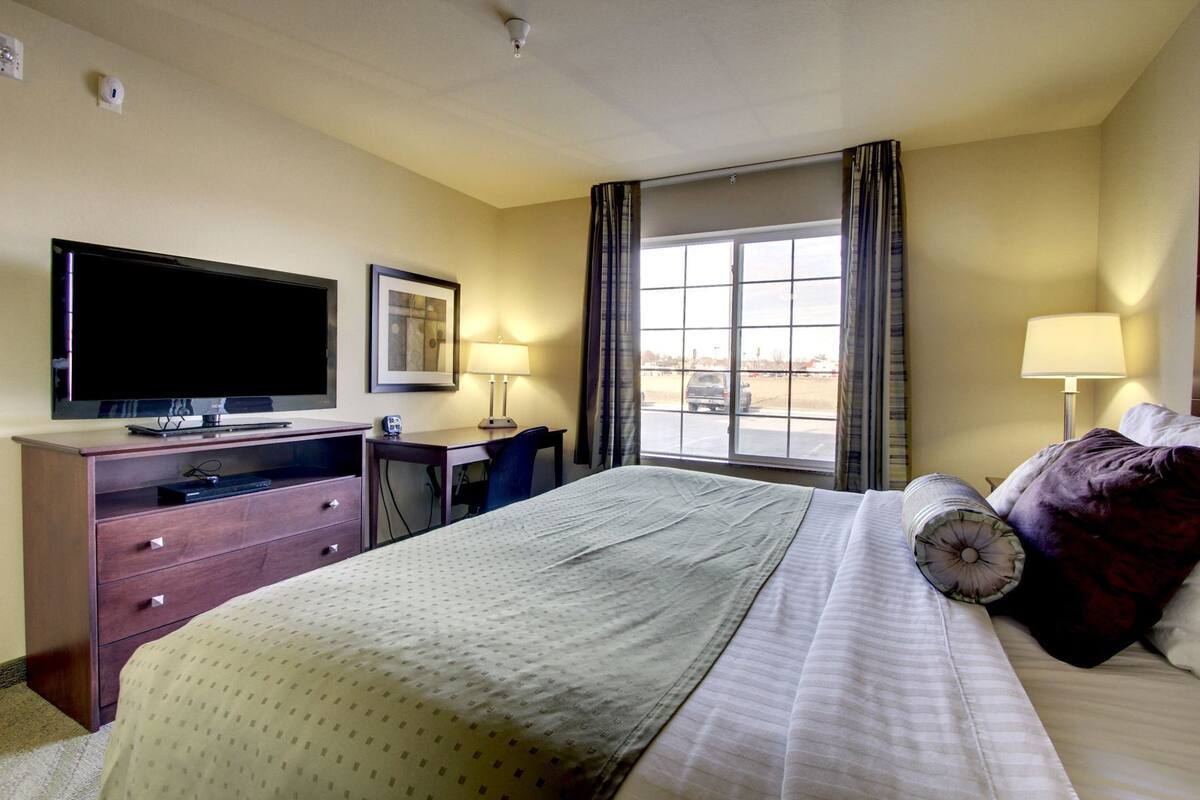 A hotel room with a bed, a desk, a television, a window with curtains, and a lamp.