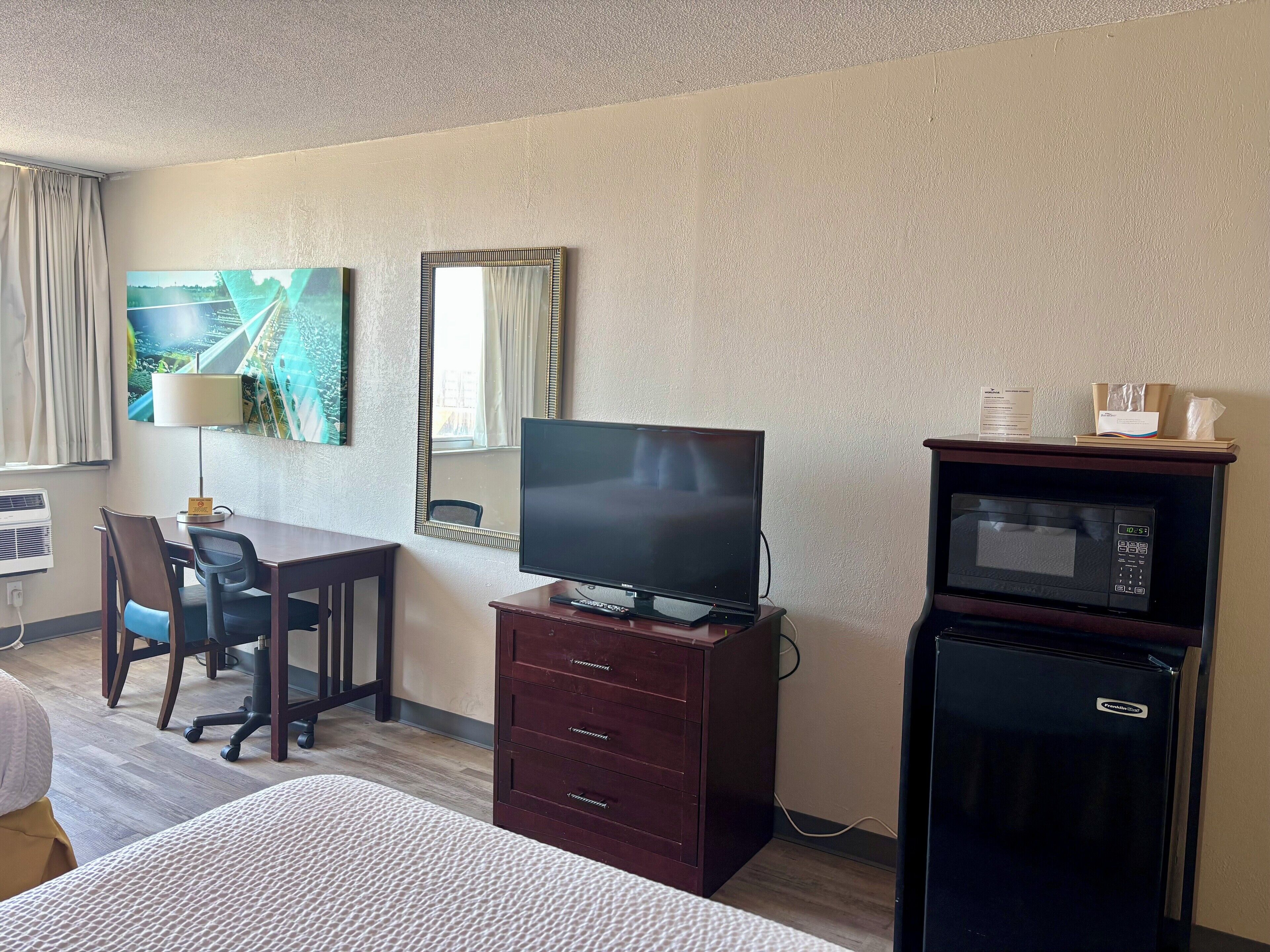 Una habitación de hotel con cama, escritorio, silla, televisor, espejo y microondas.