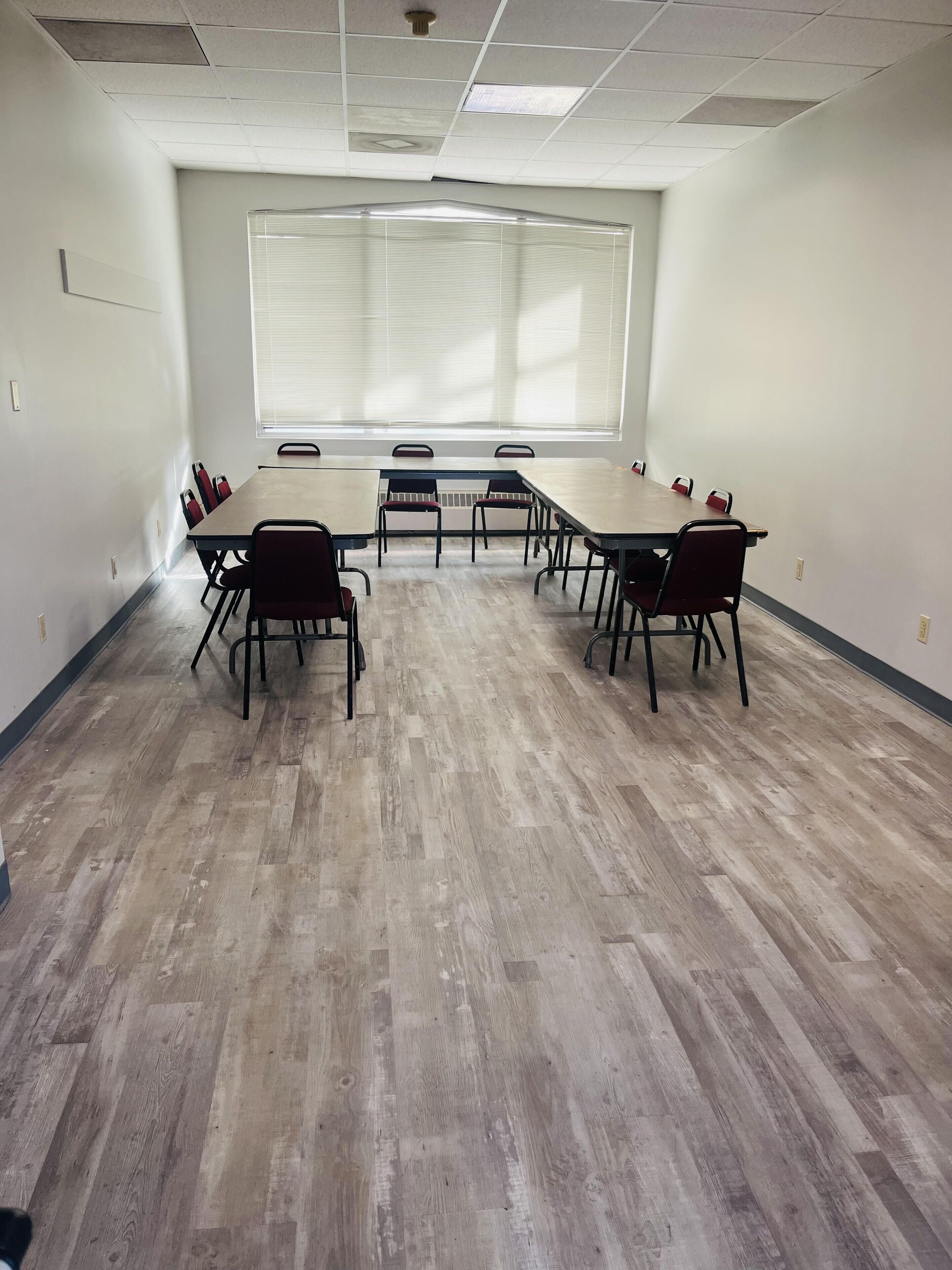 A conference room with a long table and chairs, large windows with blinds, and a wooden floor.