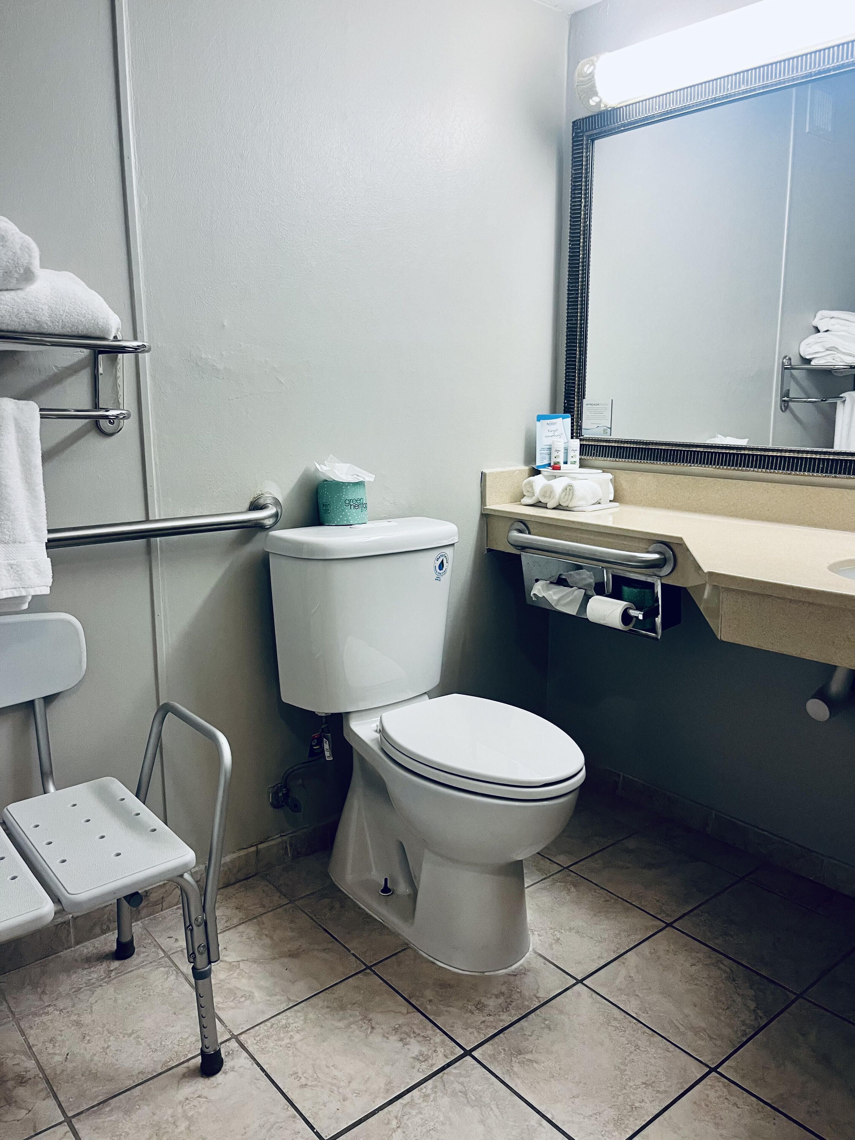 A standard hotel bathroom with a toilet, a handrail, a small step stool, a towel rack, and a mirror.