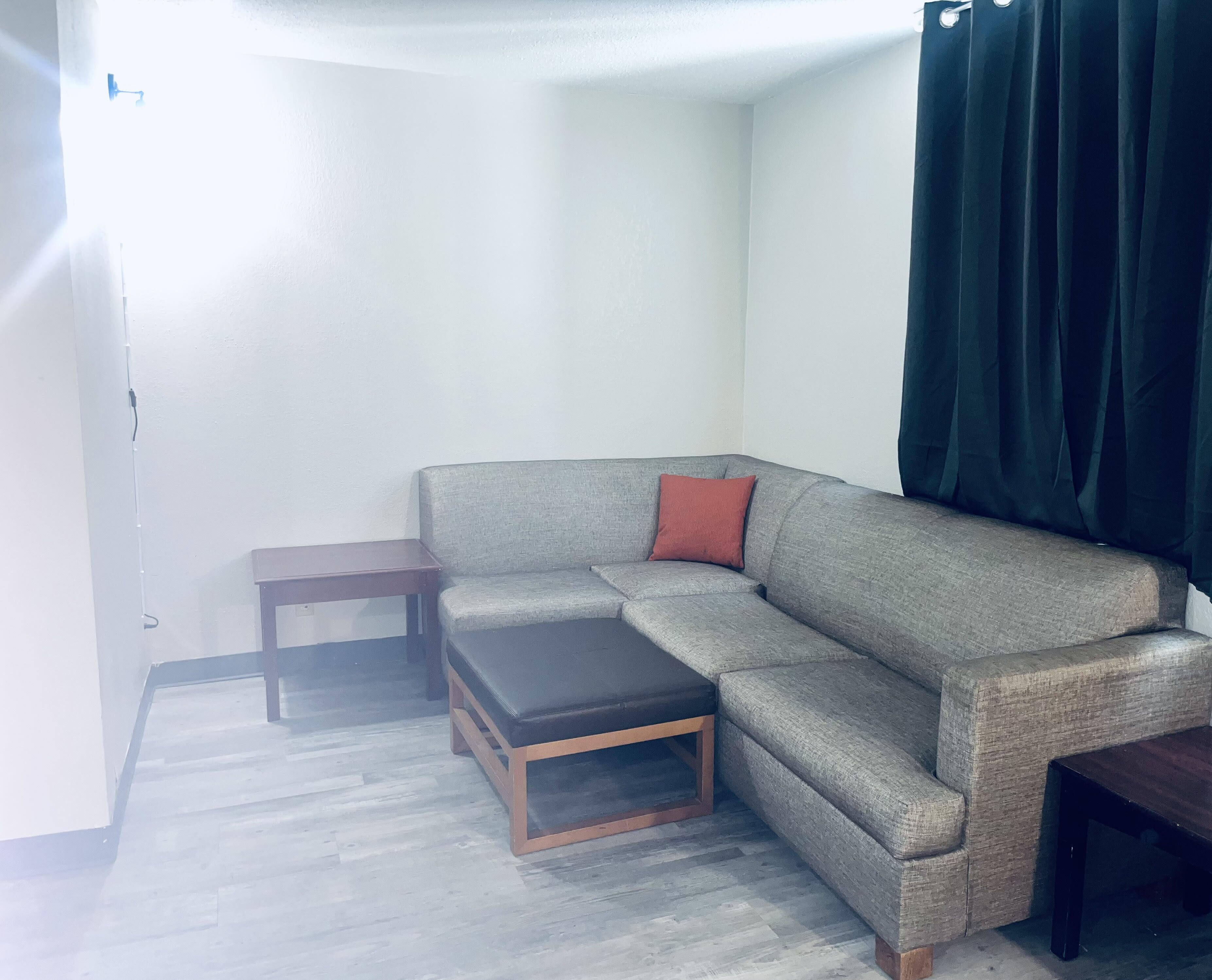 Een hoekwoonkamer met een grijze hoekbank, een klein houten bijzettafeltje en een donkerbruin poef.