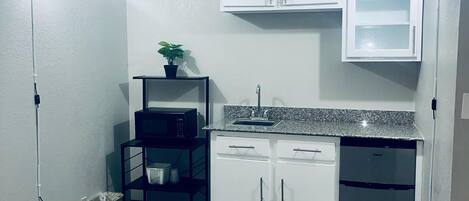 A compact kitchen with white cabinets, a granite countertop, and a dishwasher. There is a black shelf with a plant and a microwave on it.