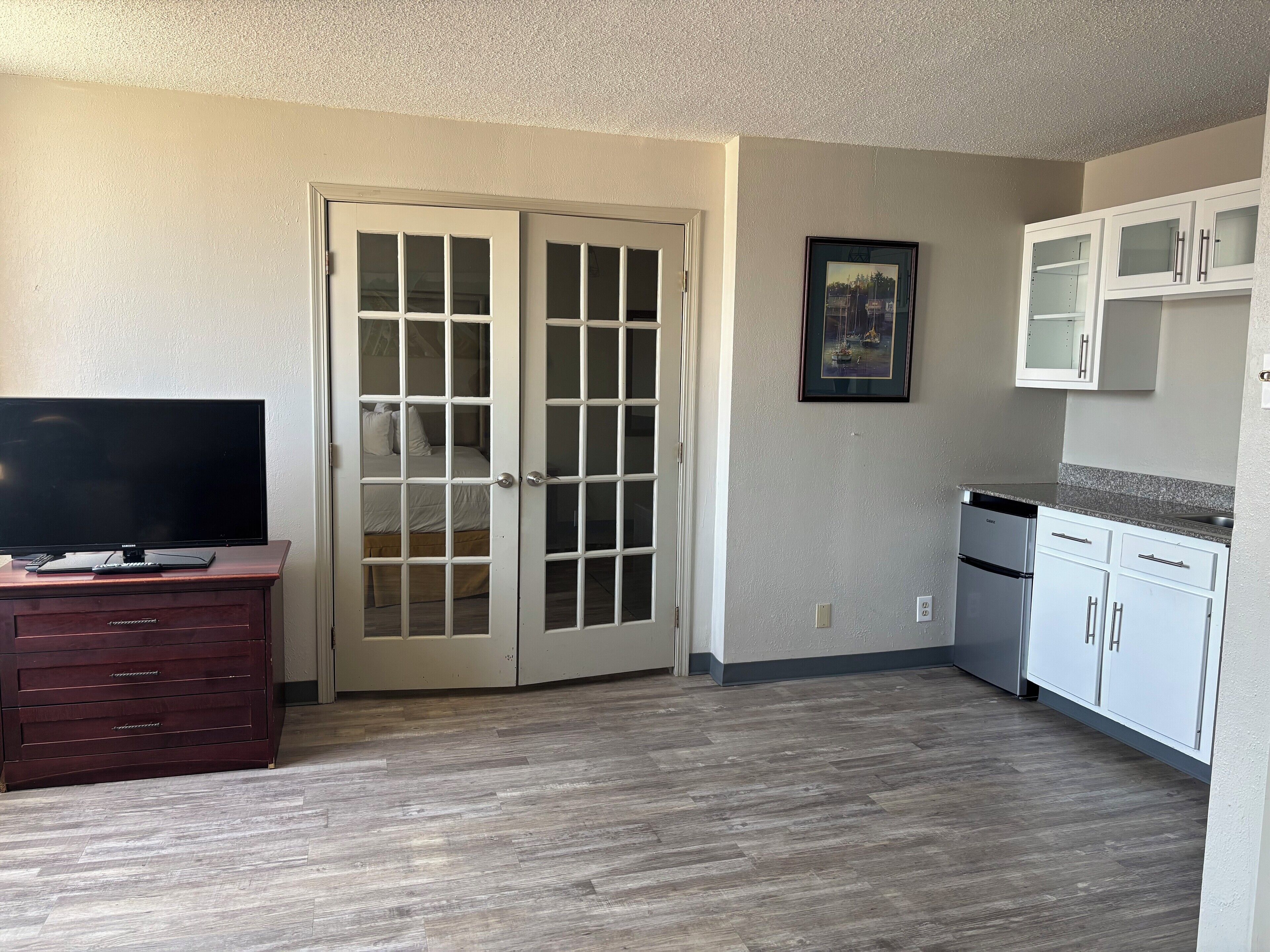 Una habitación con televisión sobre un mueble de madera, una cama con cabecera blanca, una puerta con paneles de vidrio, una zona de cocina con armarios blancos y un cuadro en la pared.