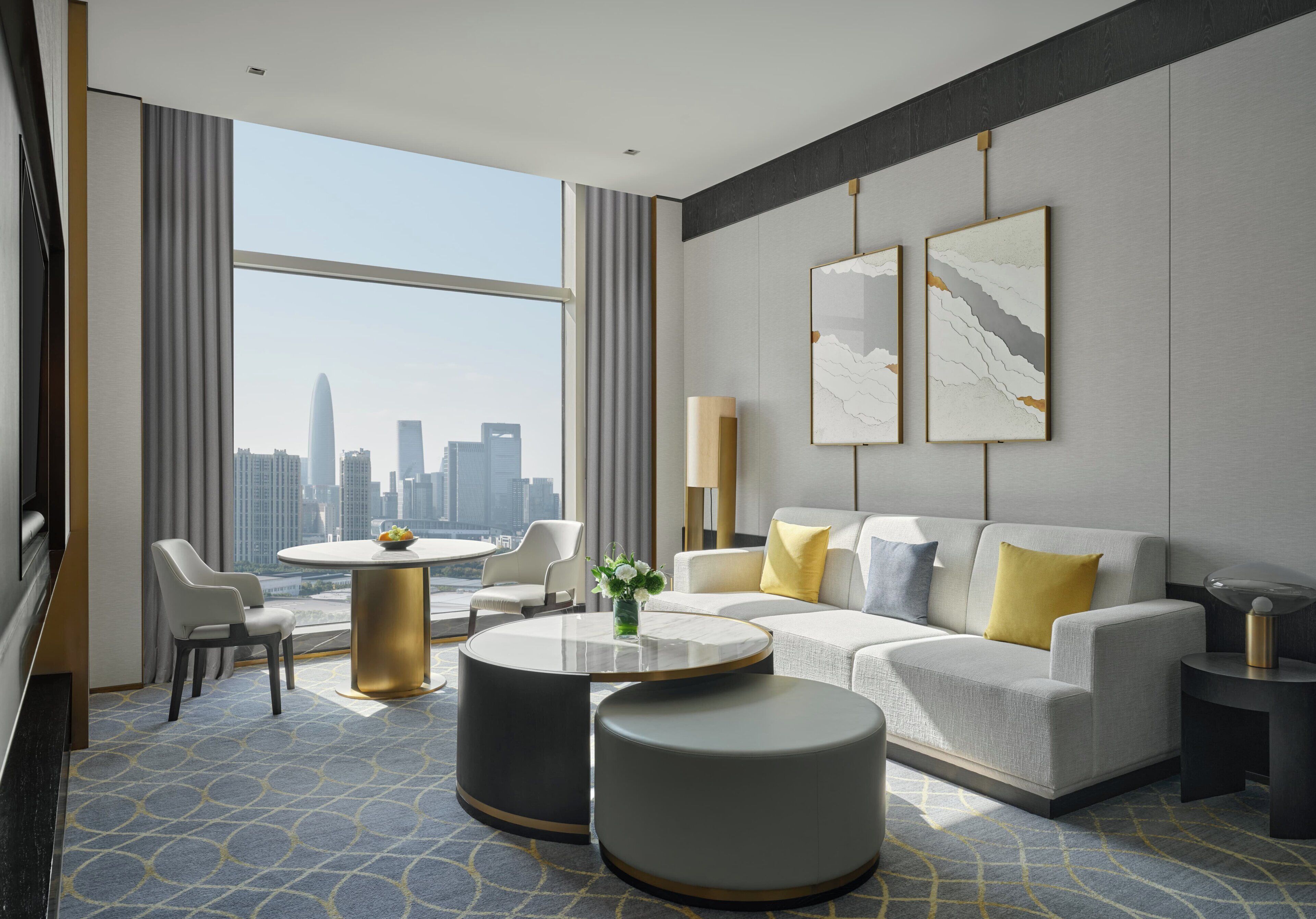 A modern hotel room with a large window, a round dining table, a sofa, and a coffee table.