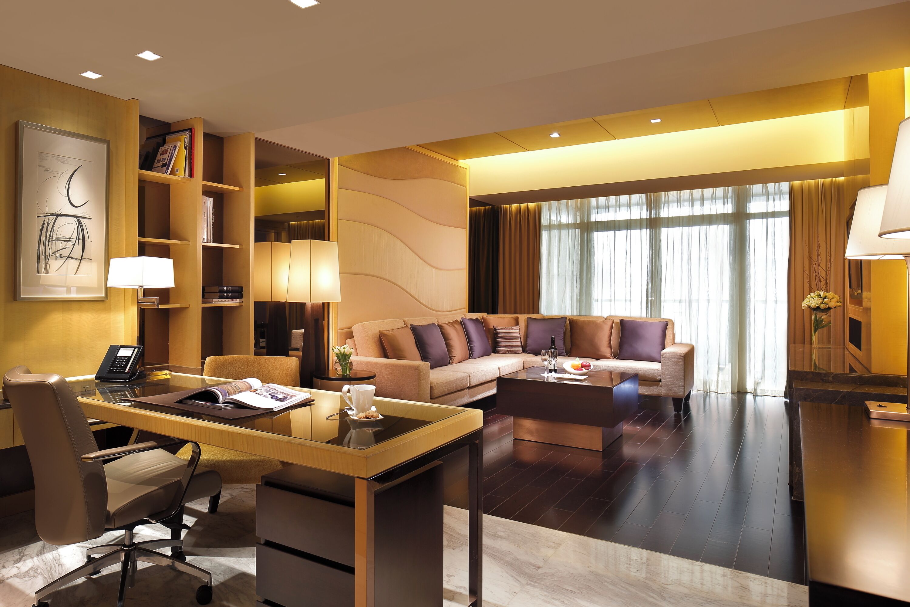 Une chambre d’hôtel moderne avec un bureau en verre, un fauteuil en cuir, un canapé et une grande fenêtre avec des rideaux.