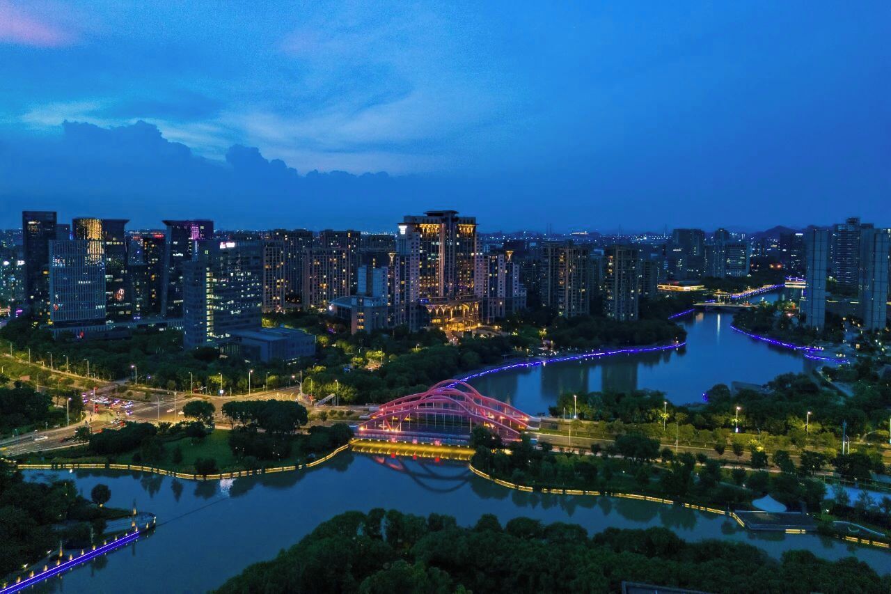 A cityscape at dusk with illuminated buildings, a river, and a bridge.