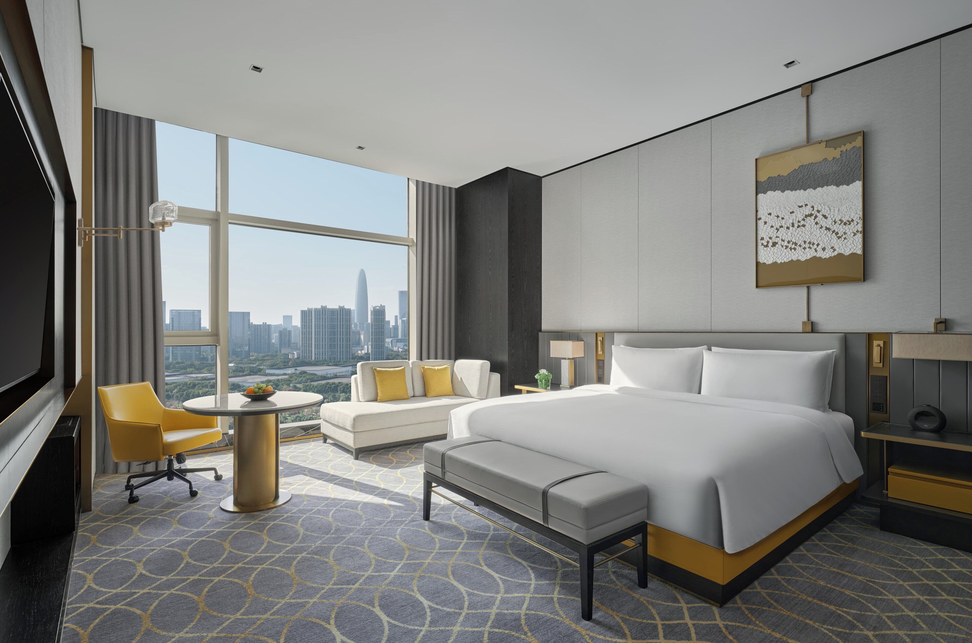 A modern hotel room with a large bed, a sofa, a desk, and a view of the city skyline.