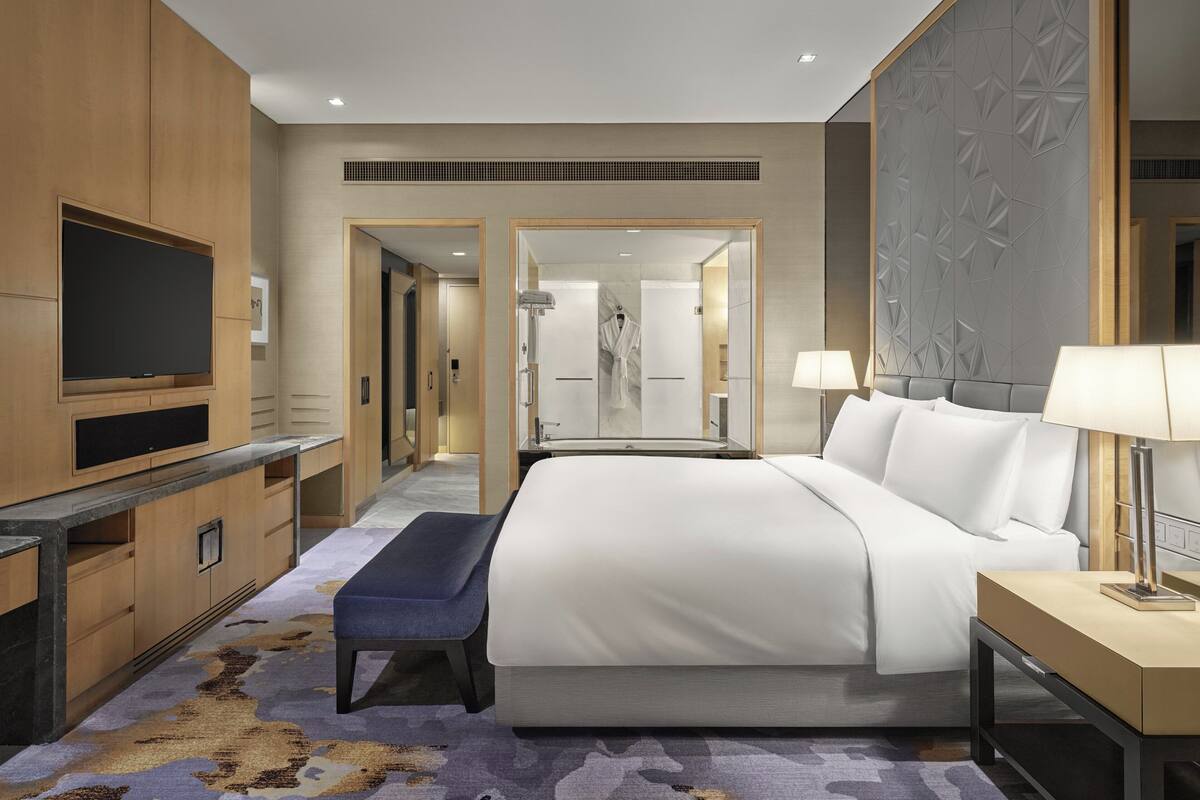 Habitación de hotel moderna con una cama grande, un televisor de pantalla plana, un banco y un baño visible a través de una puerta abierta.