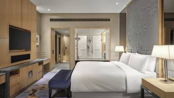 A modern hotel room with a large bed, a flat-screen TV, a bench, and a bathroom visible through an open door.