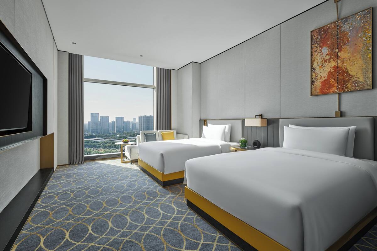 Habitación de hotel con dos camas, un televisor grande y vista a la ciudad.