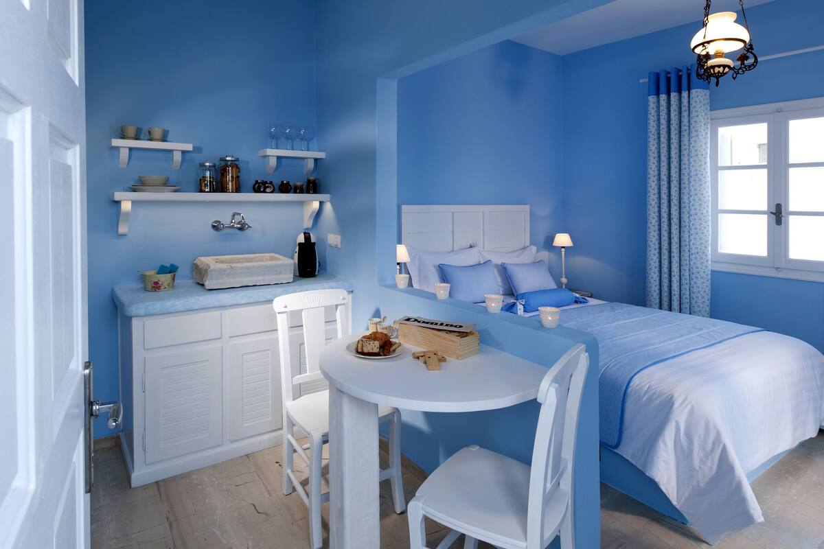 Una habitación con cama, una mesa con sillas, una pequeña cocina con estantes y una ventana con cortinas.
