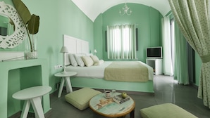 A room with a bed, a small table with a cup and book, a chair, and a television. - Senses Boutique Hotel (Santorini)
