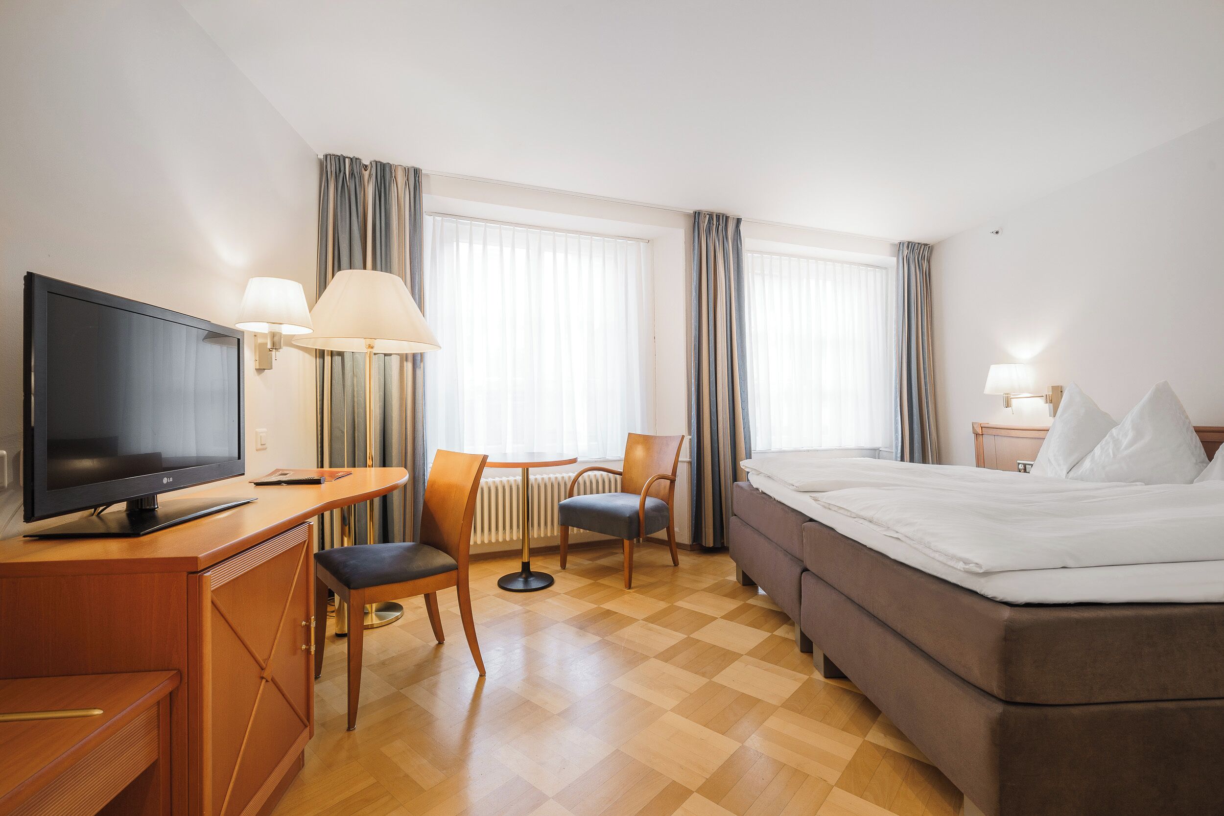 A hotel room with a bed, a TV, a desk with a chair, and a window with curtains.