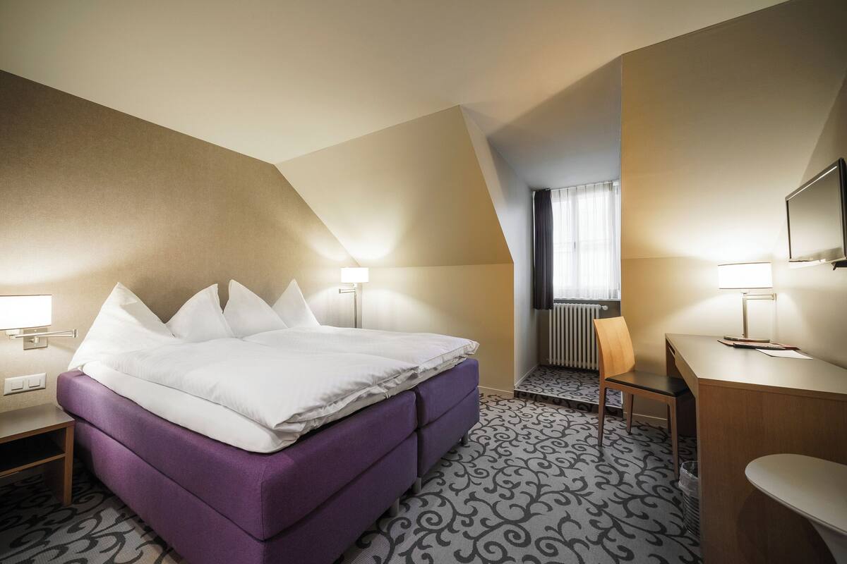 A hotel room with a large bed, a desk, a chair, and a TV.