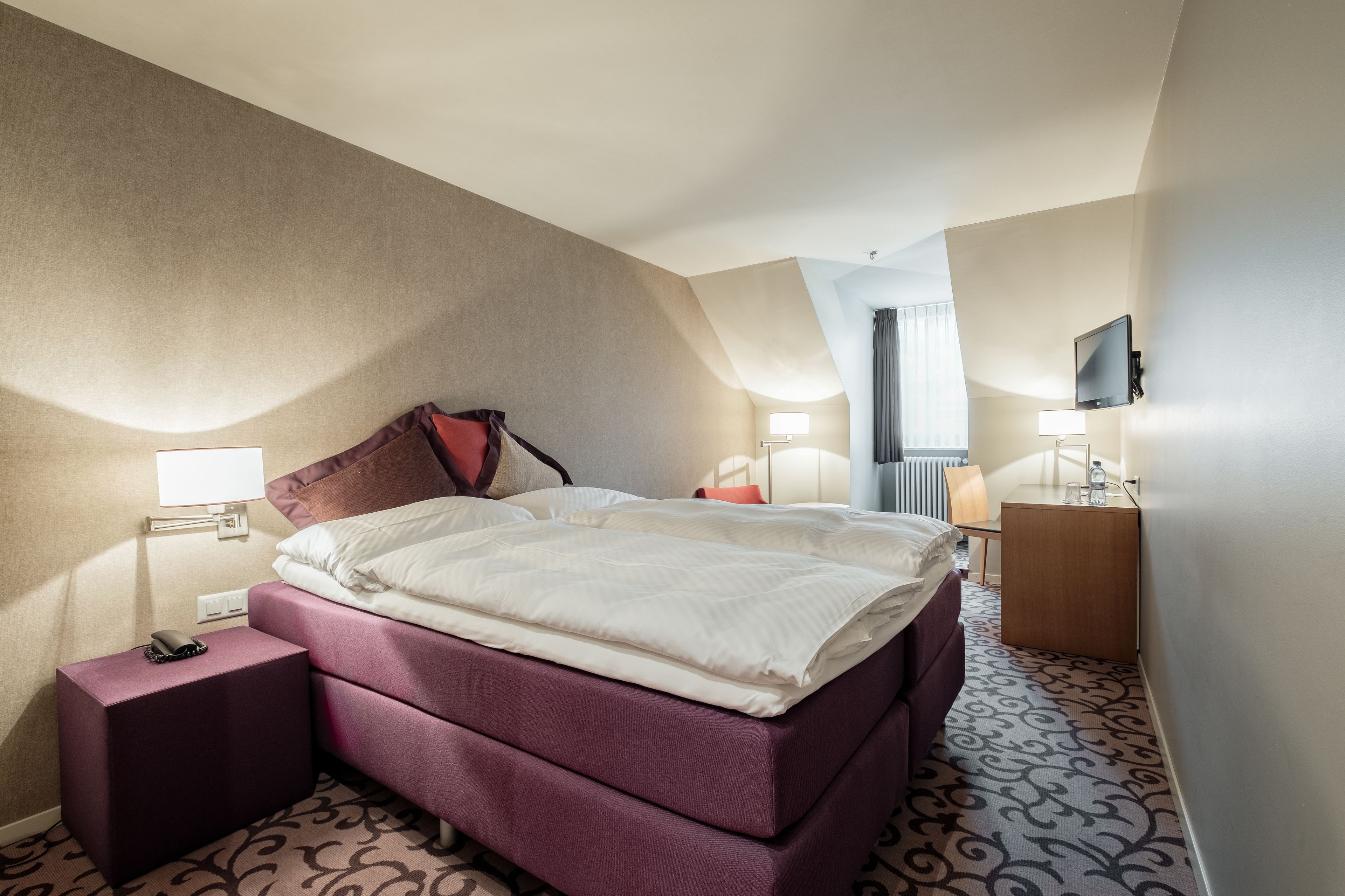 A hotel room with a large bed, a nightstand, a desk, a chair, and a television.