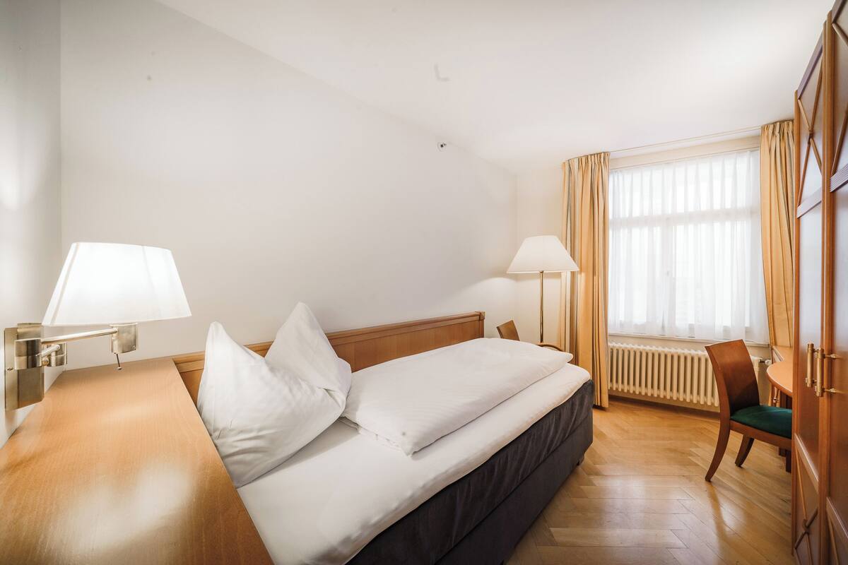 A hotel room with a bed, two bedside lamps, a chair, a radiator, and a window with curtains.