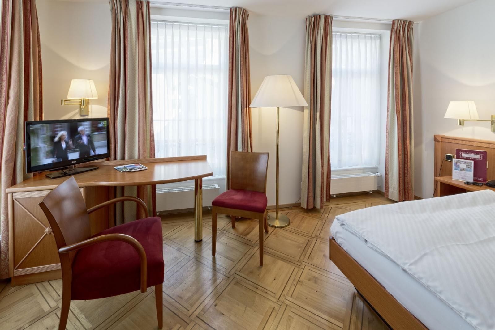 A hotel room with a bed, a desk with a television, a chair, and a floor lamp.