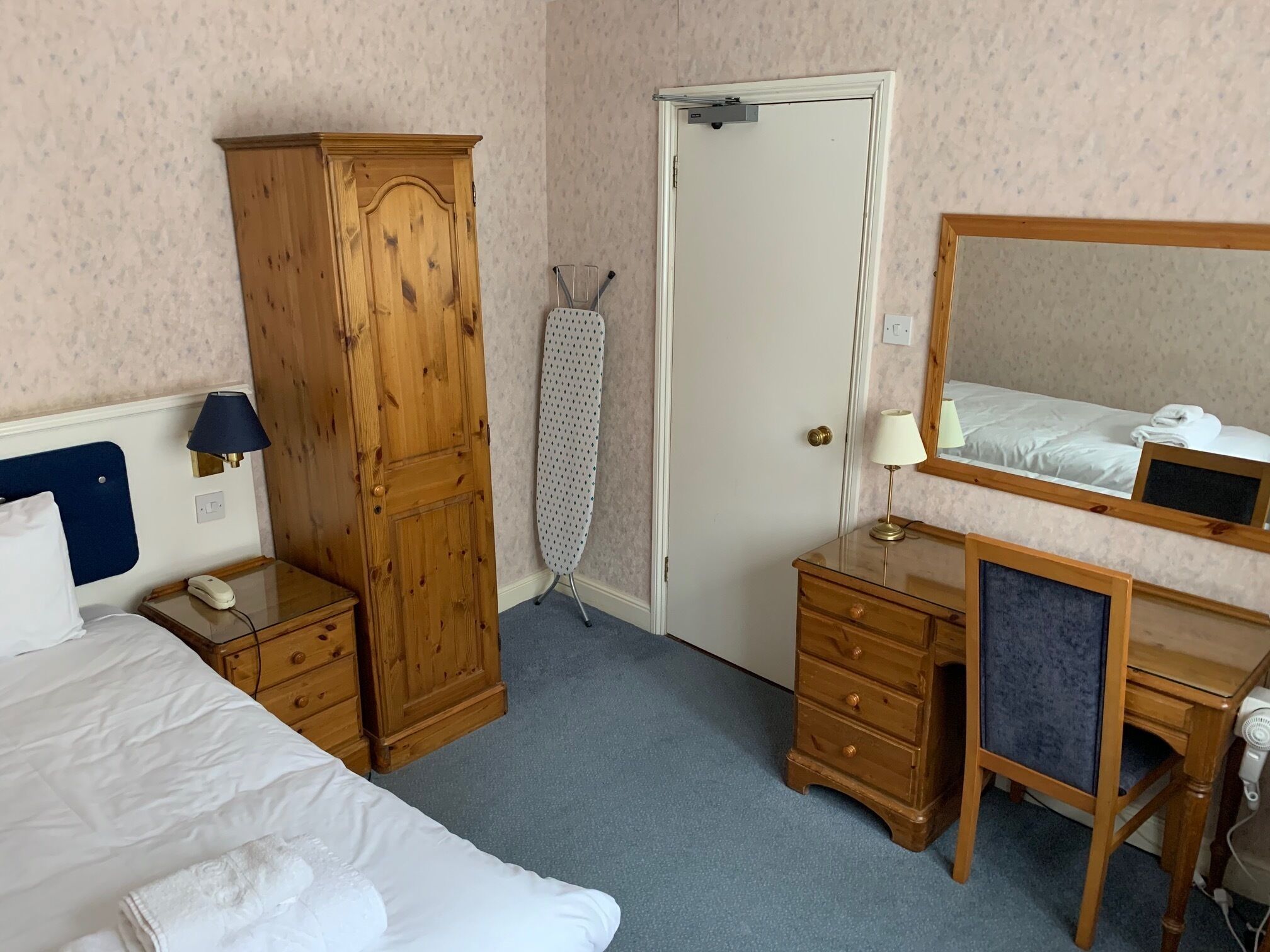A hotel room with a bed, a wooden wardrobe, a dressing table with a mirror, a small bedside table with a lamp, and a door with a towel rack.