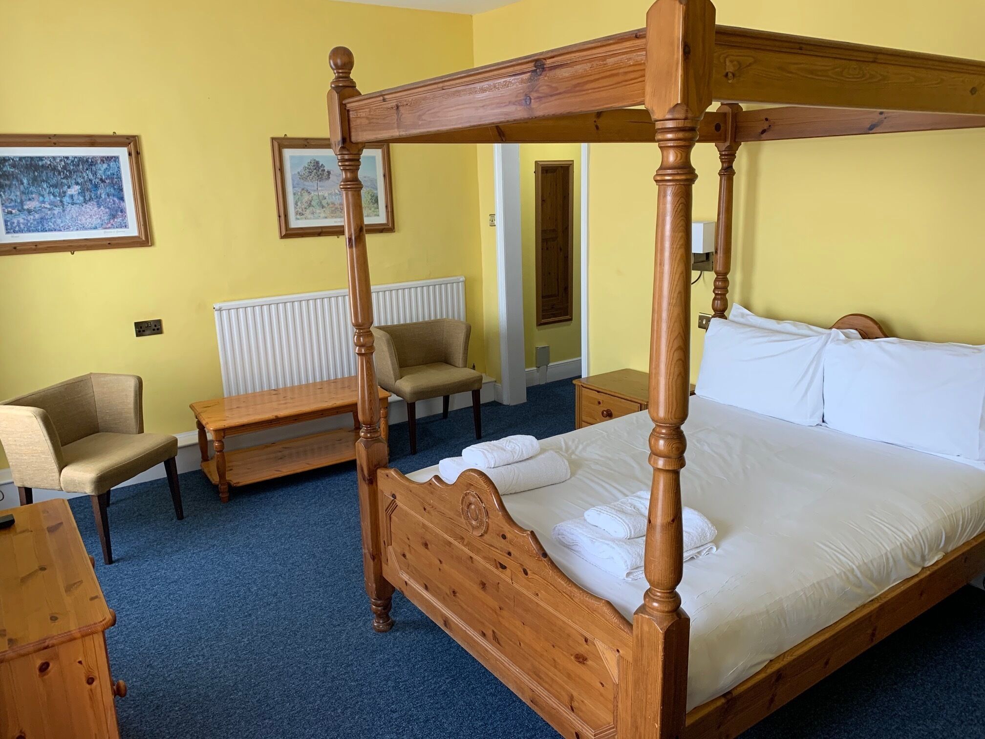 Een hemelbed met houten hoofdbord, een nachtkastje, een stoel en een bankje in een kamer met gele muren en blauw tapijt.