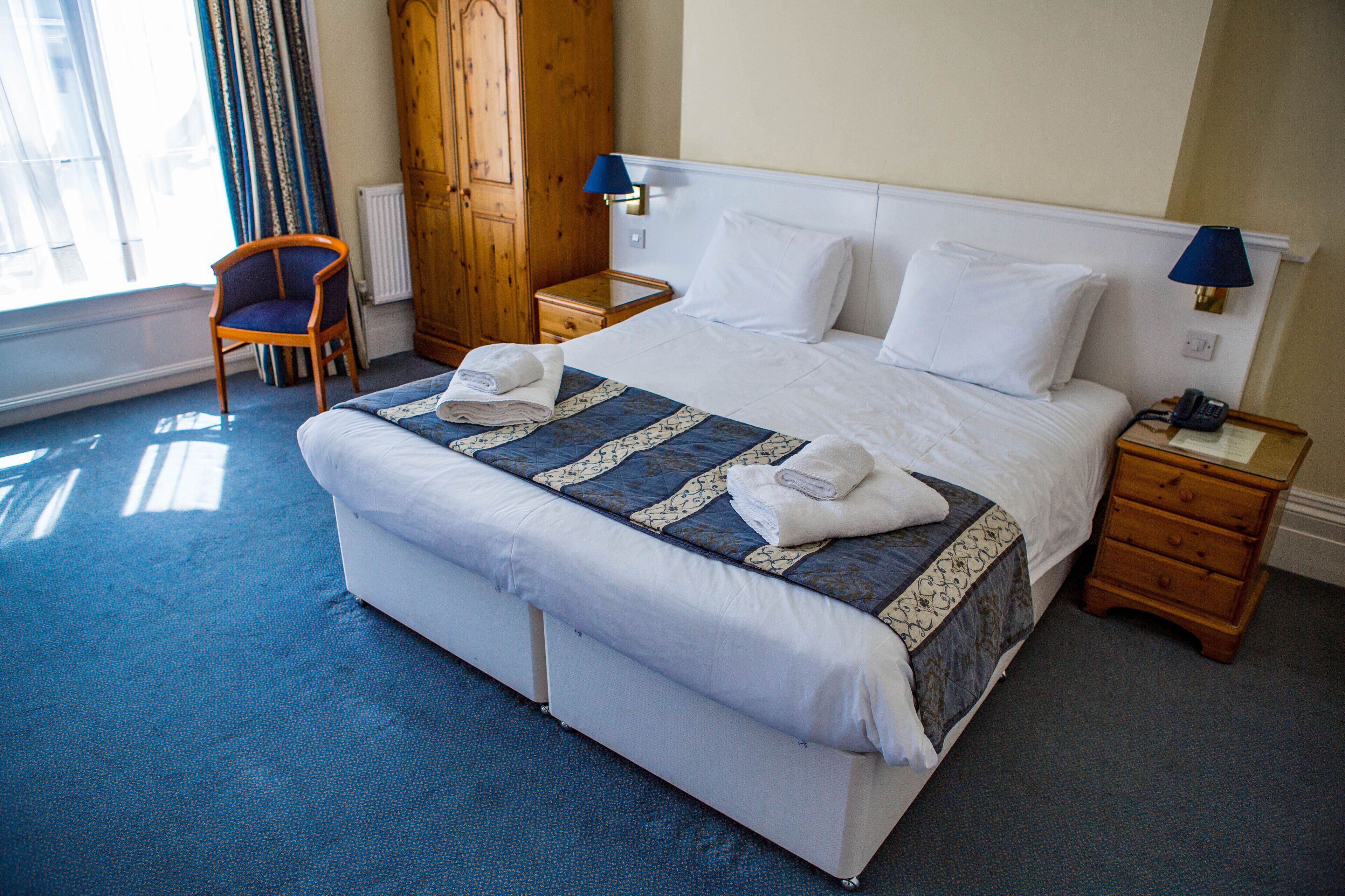 A hotel room with a large bed, a wooden wardrobe, a chair, a nightstand, and a lamp.