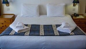 Una cama bien hecha con sábanas blancas y una colcha estampada en azul y blanco. Hay dos mesitas de noche con lámparas, un aparador de madera y un teléfono en una de las mesitas.