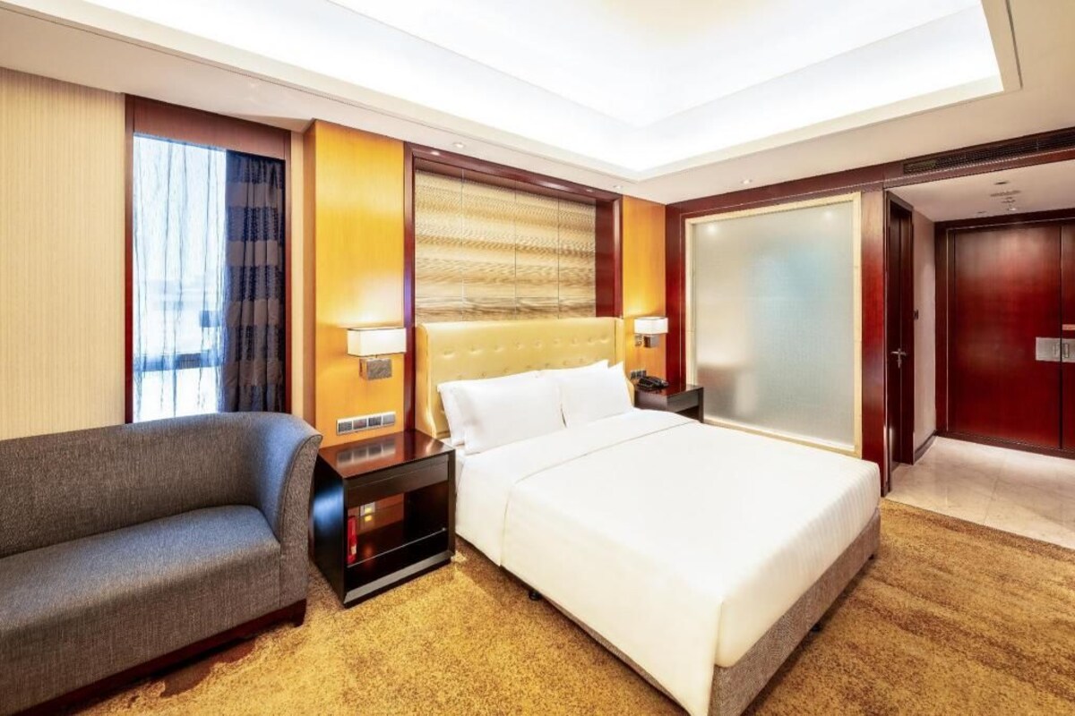 A hotel room with a bed, a sofa, a nightstand, a lamp, a sliding door, and a window with curtains.