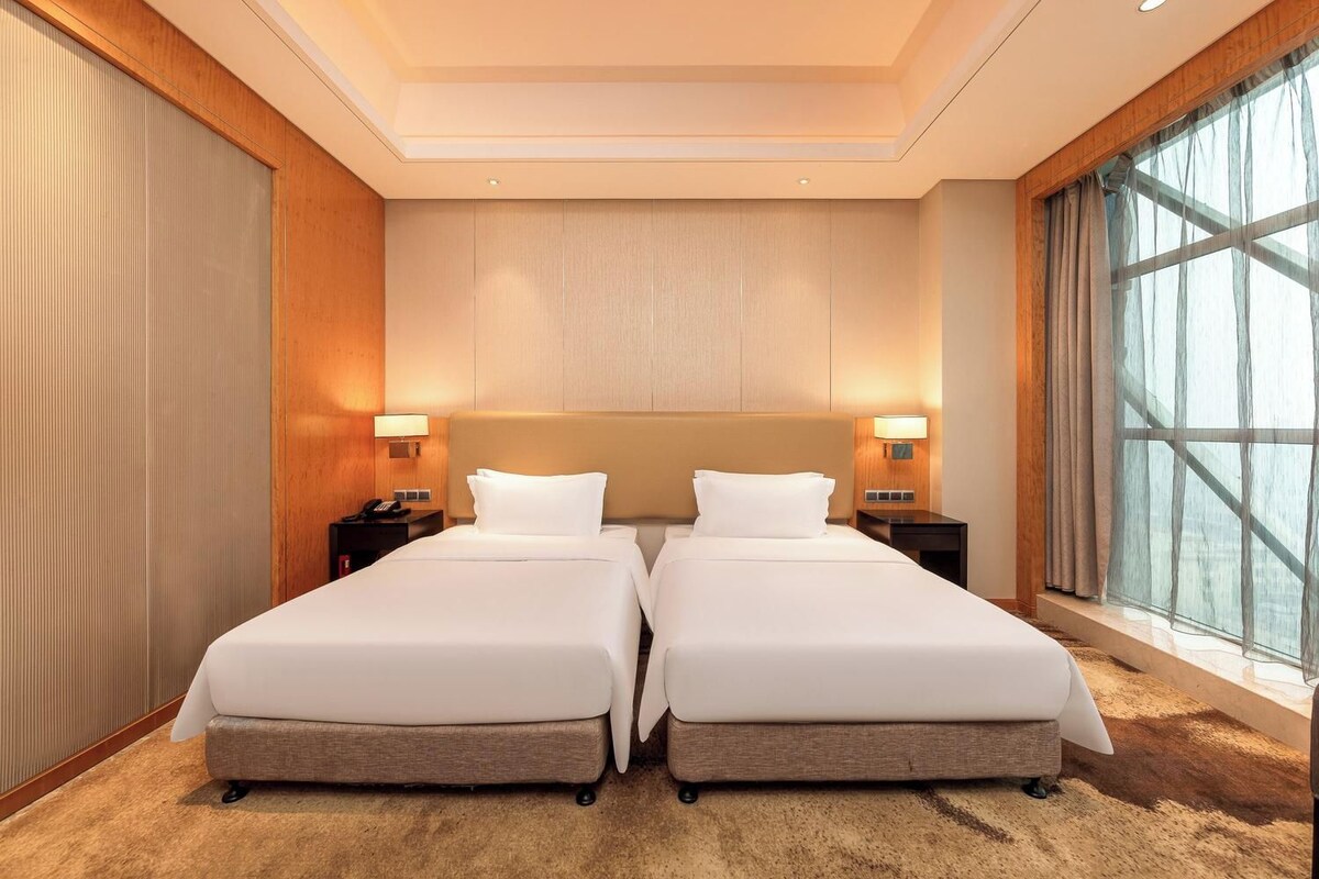 Ein Hotelzimmer mit zwei Betten, einem großen Fenster mit Vorhängen und einem Teppichboden.