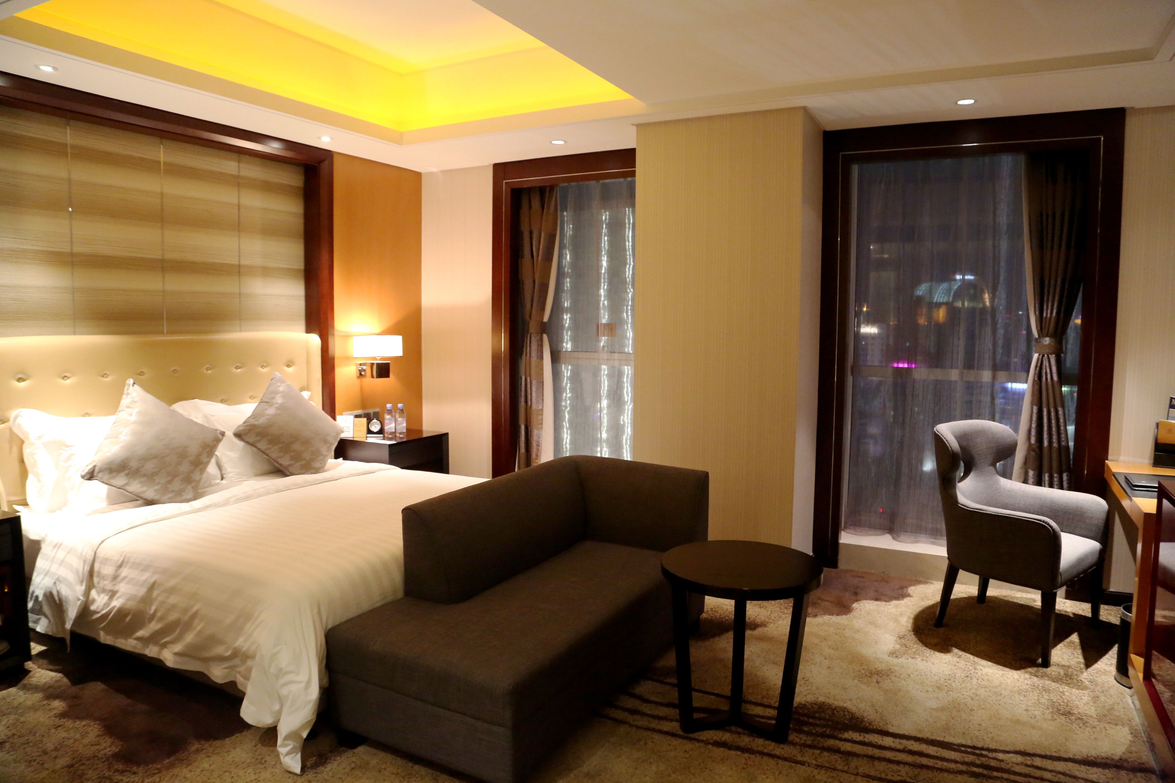 A hotel room with a bed, a sofa, a chair, a small table, and a large window with curtains.