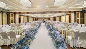 Un salón de banquetes con mesas redondas, sillas blancas y un pasillo decorado con flores.