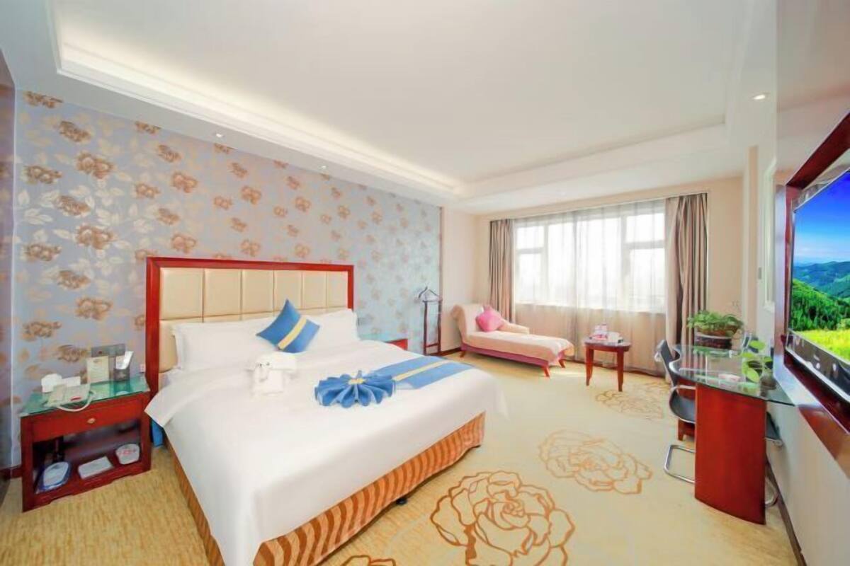 Une chambre d’hôtel avec un grand lit, un bureau avec une télévision, une fenêtre donnant sur un beau paysage et un petit balcon.
