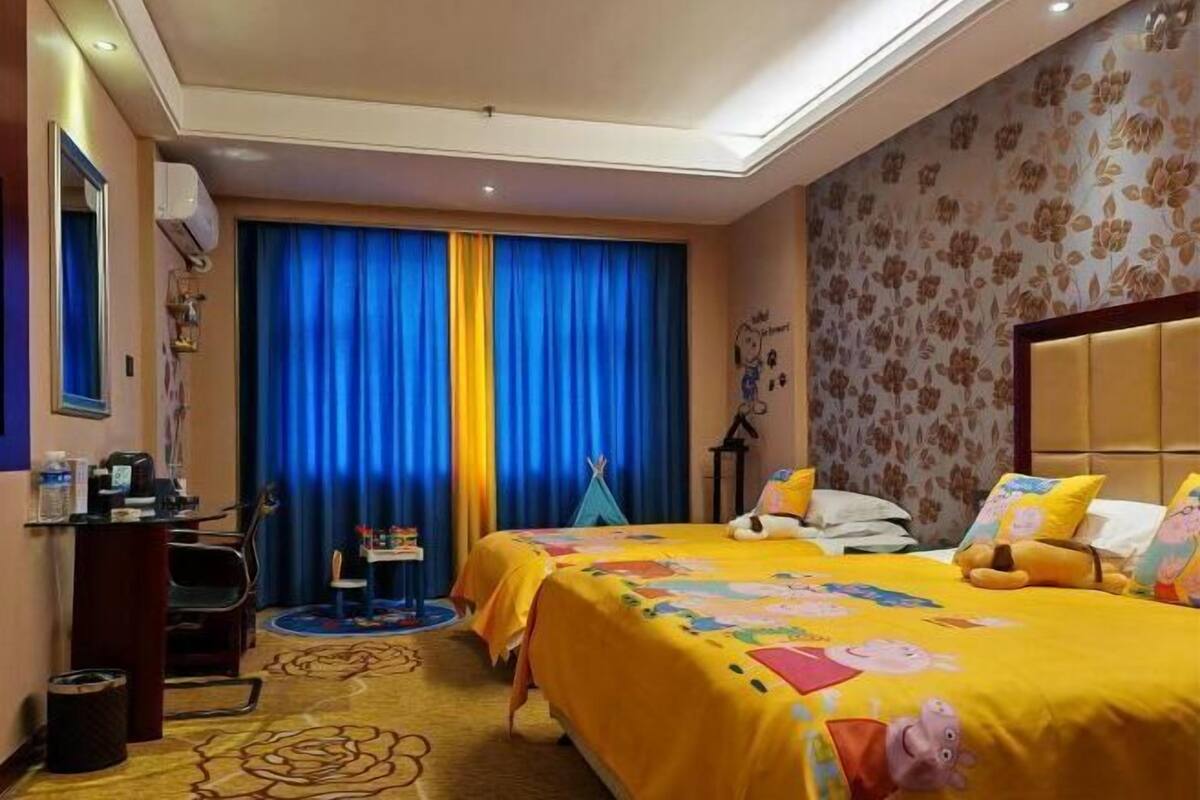 Une chambre d’hôtel avec deux lits, un bureau, une chaise, une fenêtre avec des rideaux bleus et un motif floral sur le mur.