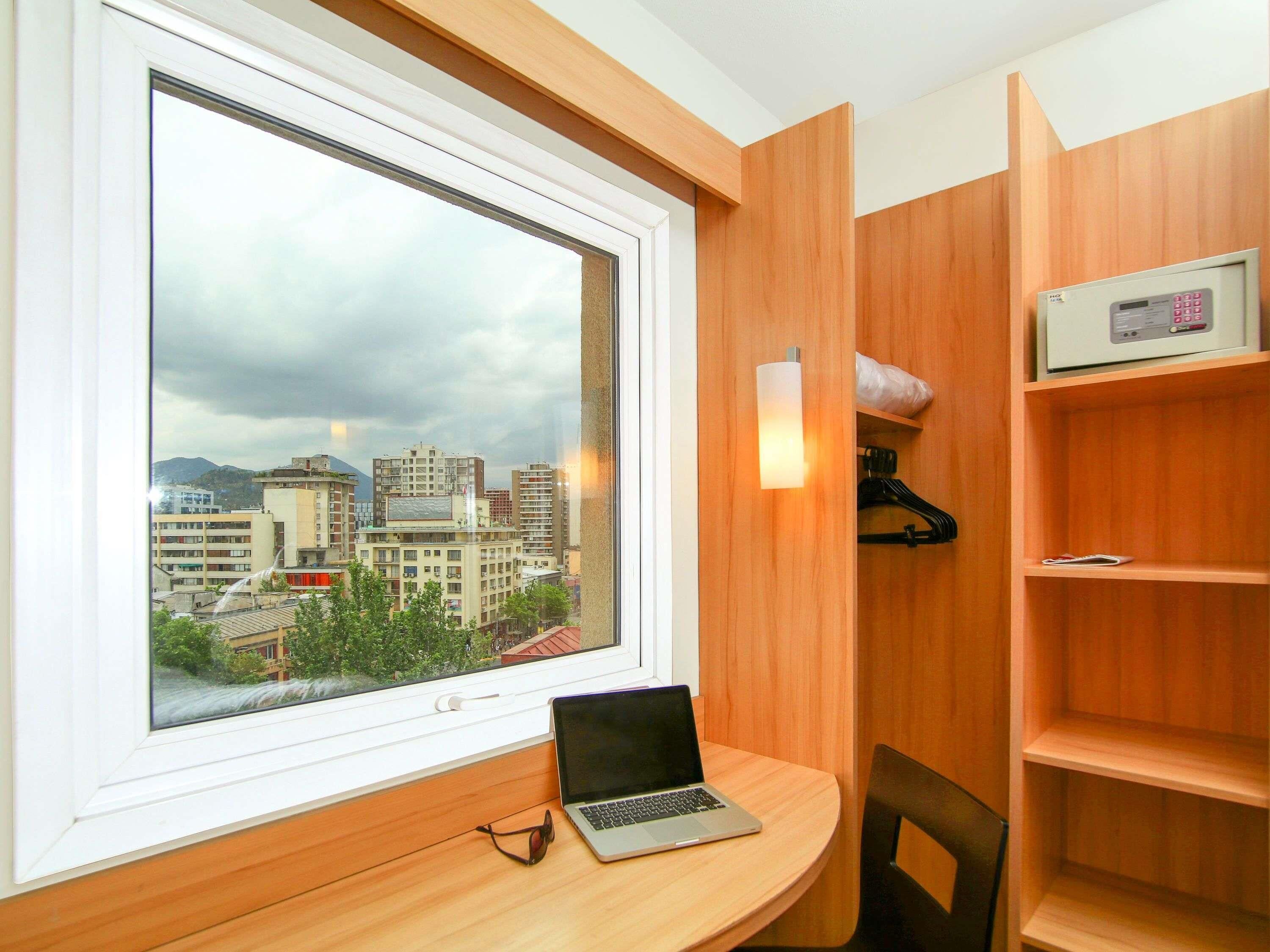 A room with a large window offering a city view, a wooden desk with a laptop, a chair, and a shelf with a clock.
