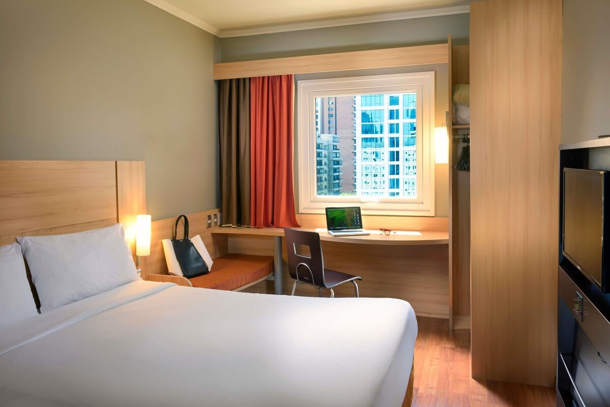 Een hotelkamer met een bed, een bureau met een laptop, een stoel, een televisie en een raam met gordijnen.