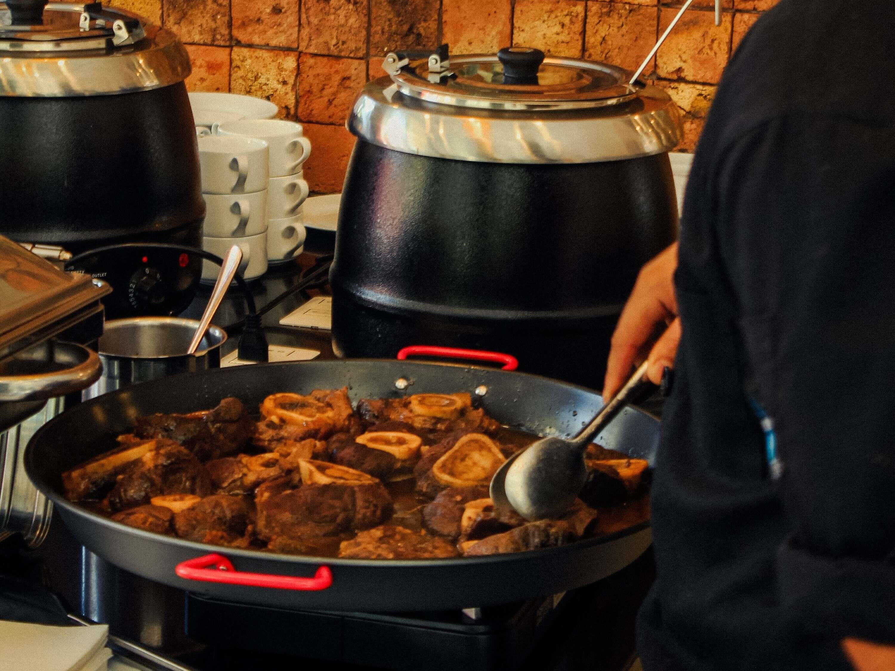 A kitchen with a large pan of meat being cooked, a stove, and a pot on the counter.