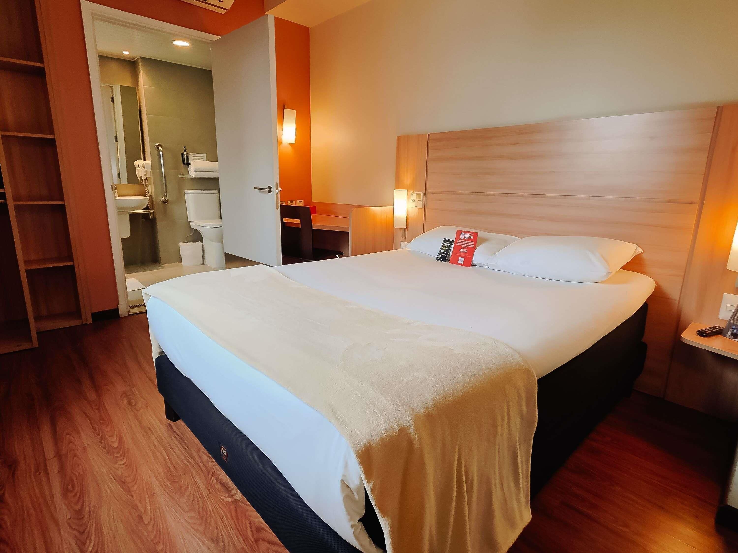 A hotel room with a large bed, a wooden headboard, a bedside table with a lamp, and a bathroom visible through an open door.