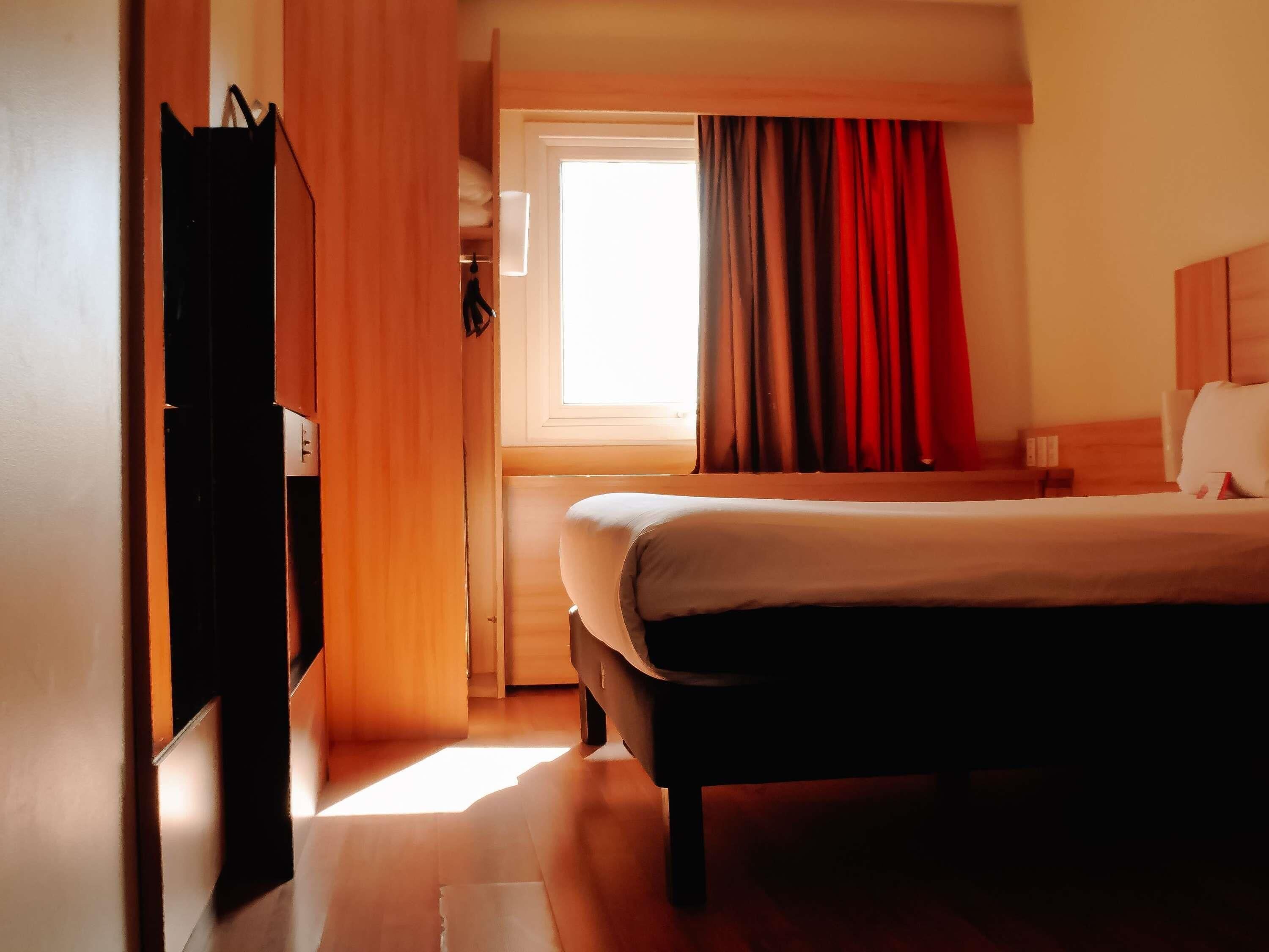 Una habitación de hotel con cama, armario, ventana con cortinas y mesita de noche.