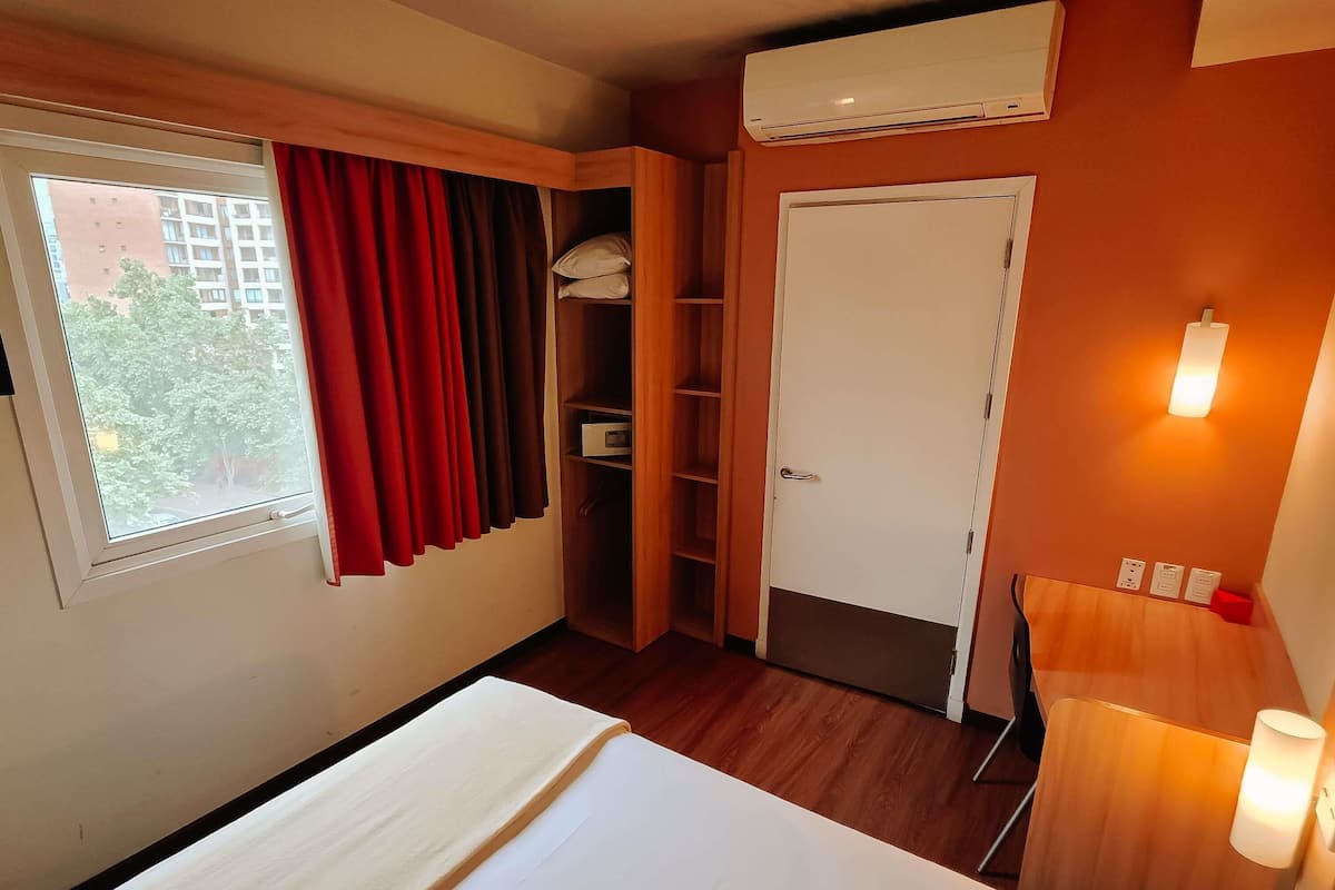 A hotel room with a bed, a wardrobe, a door, an air conditioning unit, and a window with curtains.