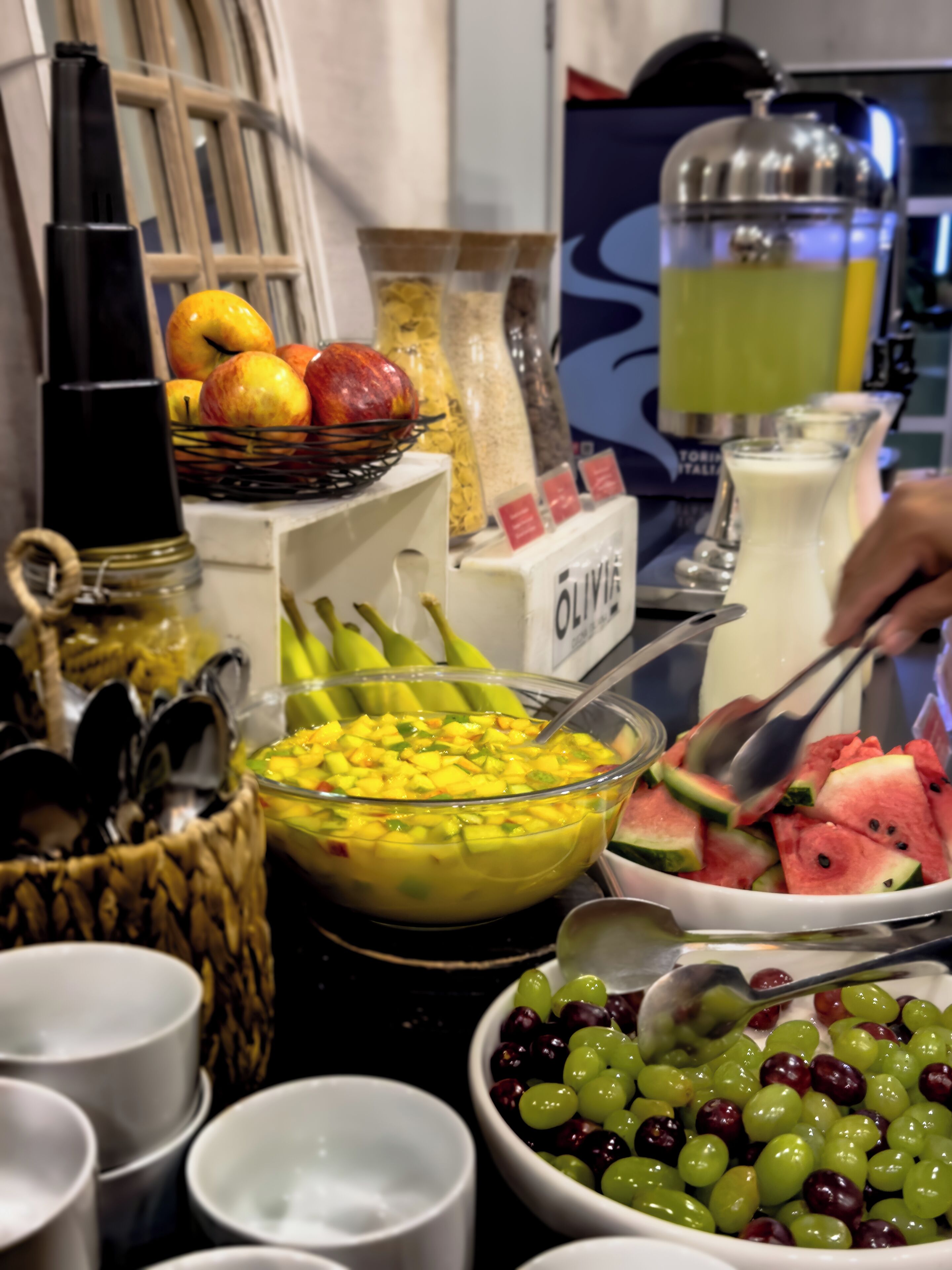 Un bufé con diversos alimentos, incluyendo frutas, un tazón de ensalada de mango y un tazón de uvas.
