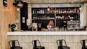 Un espace bar avec un comptoir, des étagères remplies de bouteilles, une machine à café et trois tabourets noirs.