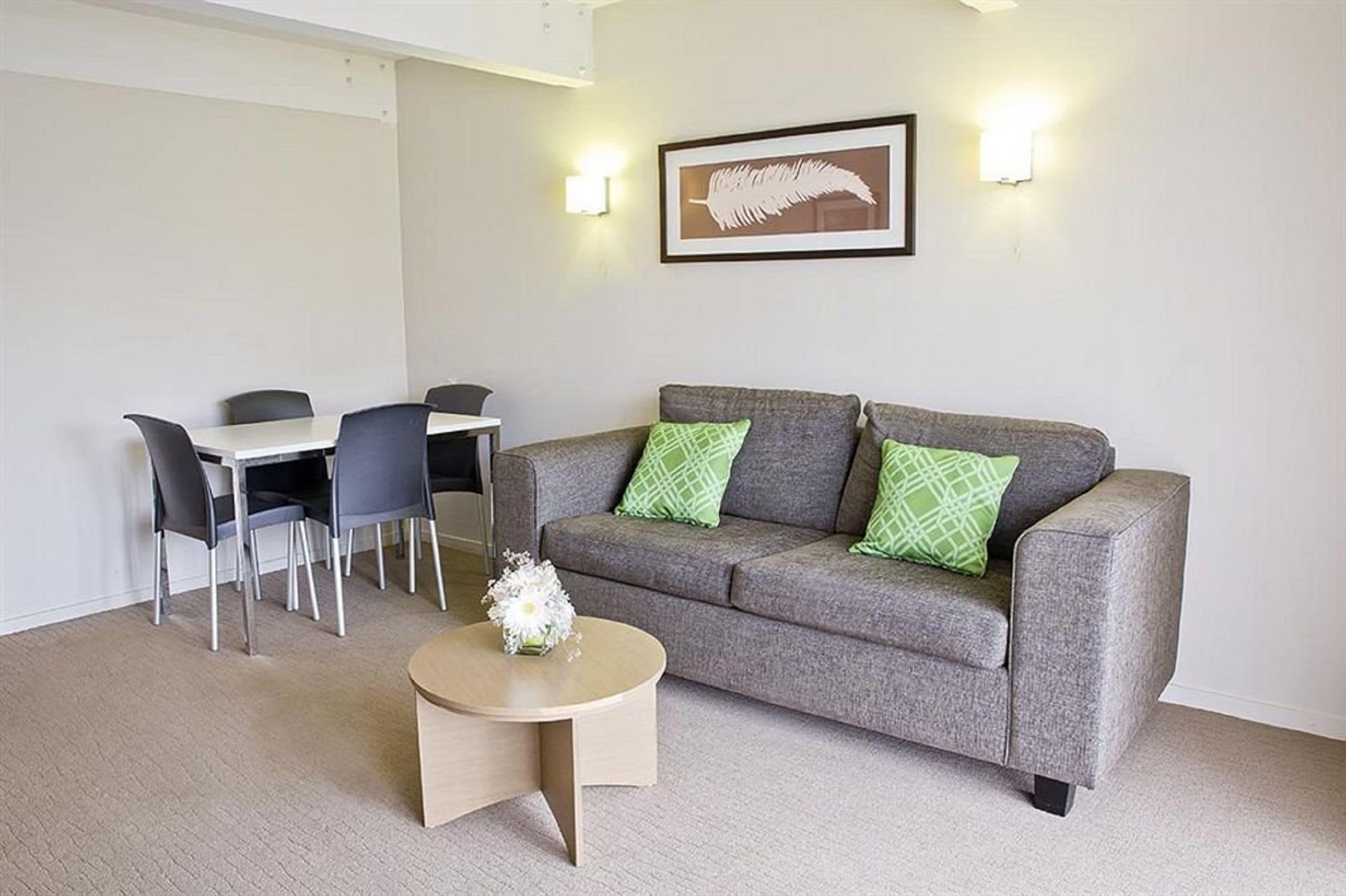A modern living room with a grey sofa, two green pillows, a round coffee table with a white flower, a dining table with chairs, and a framed artwork on the wall.
