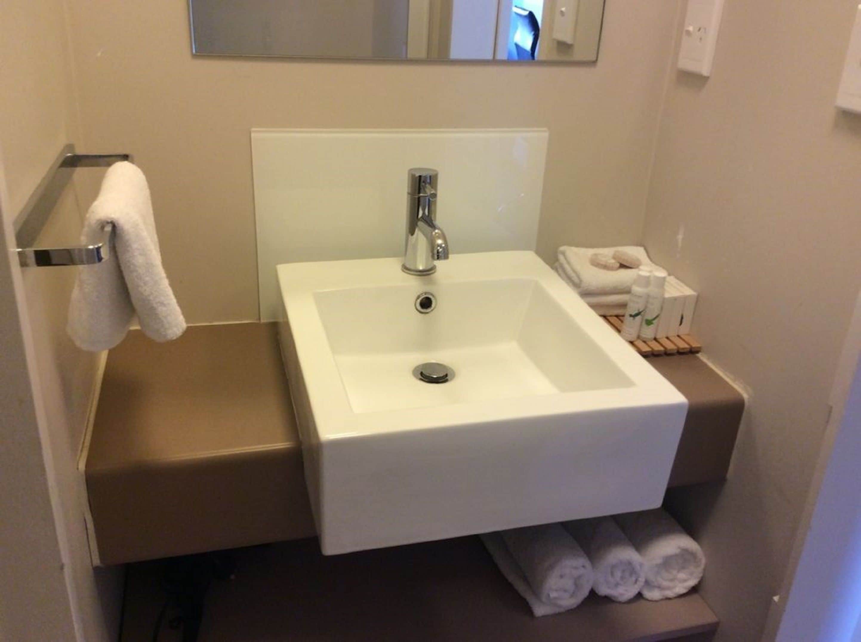 Une salle de bain avec un lavabo blanc, un miroir au-dessus et un porte-serviettes avec une serviette. On y trouve également des serviettes et des articles de toilette sur une étagère.