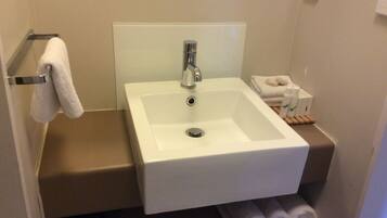 Une salle de bain avec un lavabo blanc, un miroir au-dessus et un porte-serviettes avec une serviette. On y trouve également des serviettes et des articles de toilette sur une étagère.