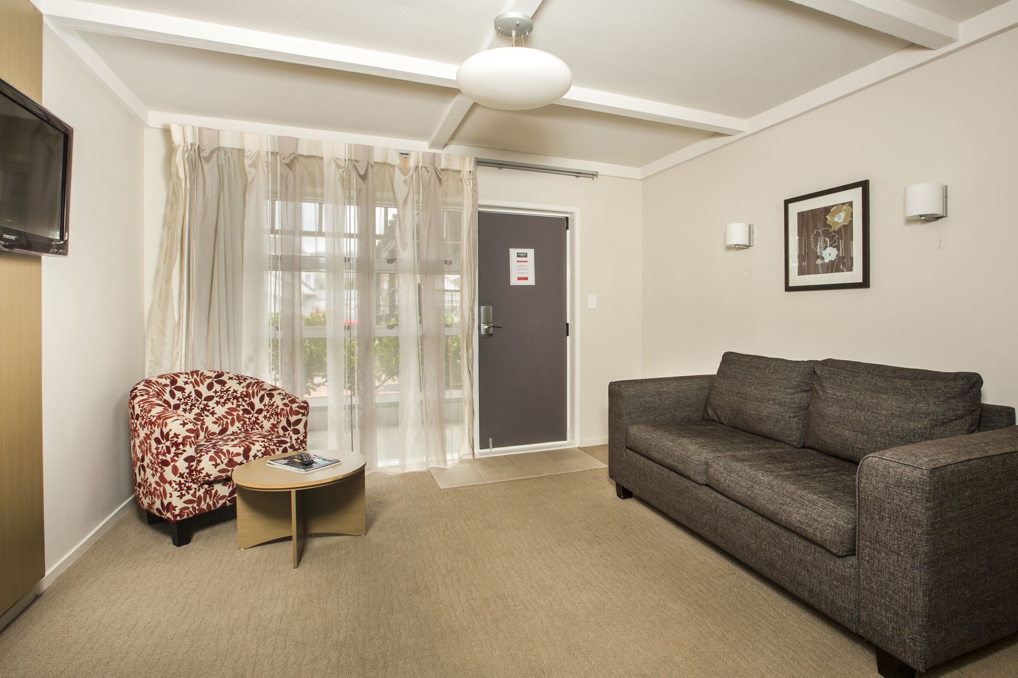 Ein Zimmer mit grauem Sofa, einem kleinen runden Tisch, einem an der Wand befestigten Fernseher, einer Tür mit einem Schild und einem Fenster mit durchscheinenden Vorhängen.