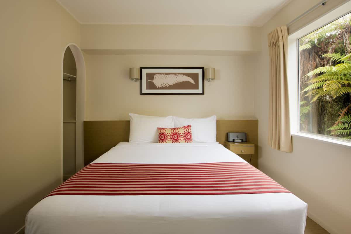 Quarto de hotel com cama, uma obra de arte emoldurada na parede, mesa de cabeceira e janela com cortinas.